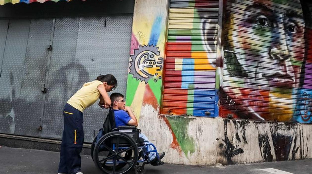 Una mujer empuja a un hombre en silla de ruedas frente a un graffiti del héroe nacional Simón Bolívar.